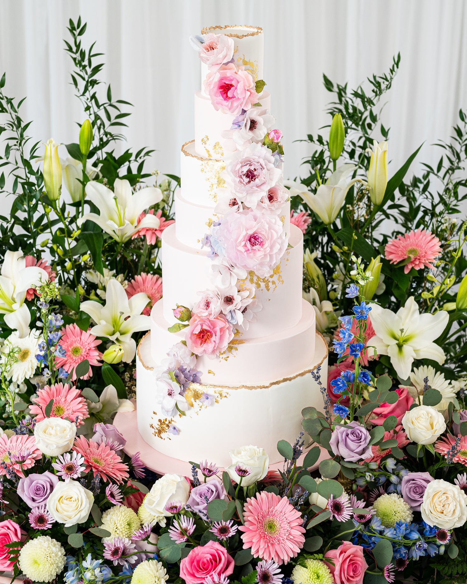 editorial tall wedding cake by sugar nursery with handmade wafer paper flowers in pink, blue, and lavender, gold leaf accents, live edge cake surrounded by flowers